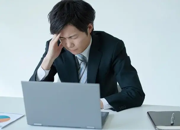 パソコンを前に頭に手を当てて悩んでいる人の画像